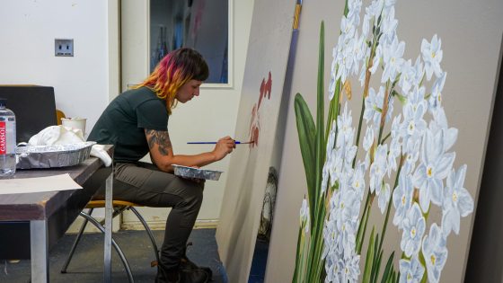 A person in an art studio hunched over painting flowers on a large-scale white board.